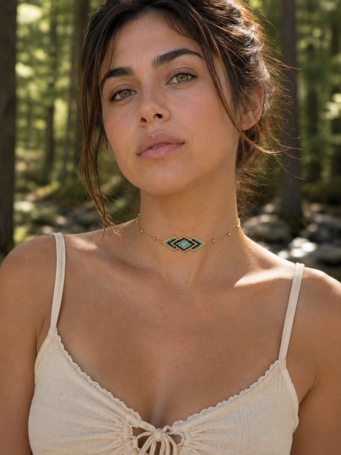 A woman wearing a gold geometric chevron necklace with turquoise and white accents, photographed outdoors in a sunlit forest setting.