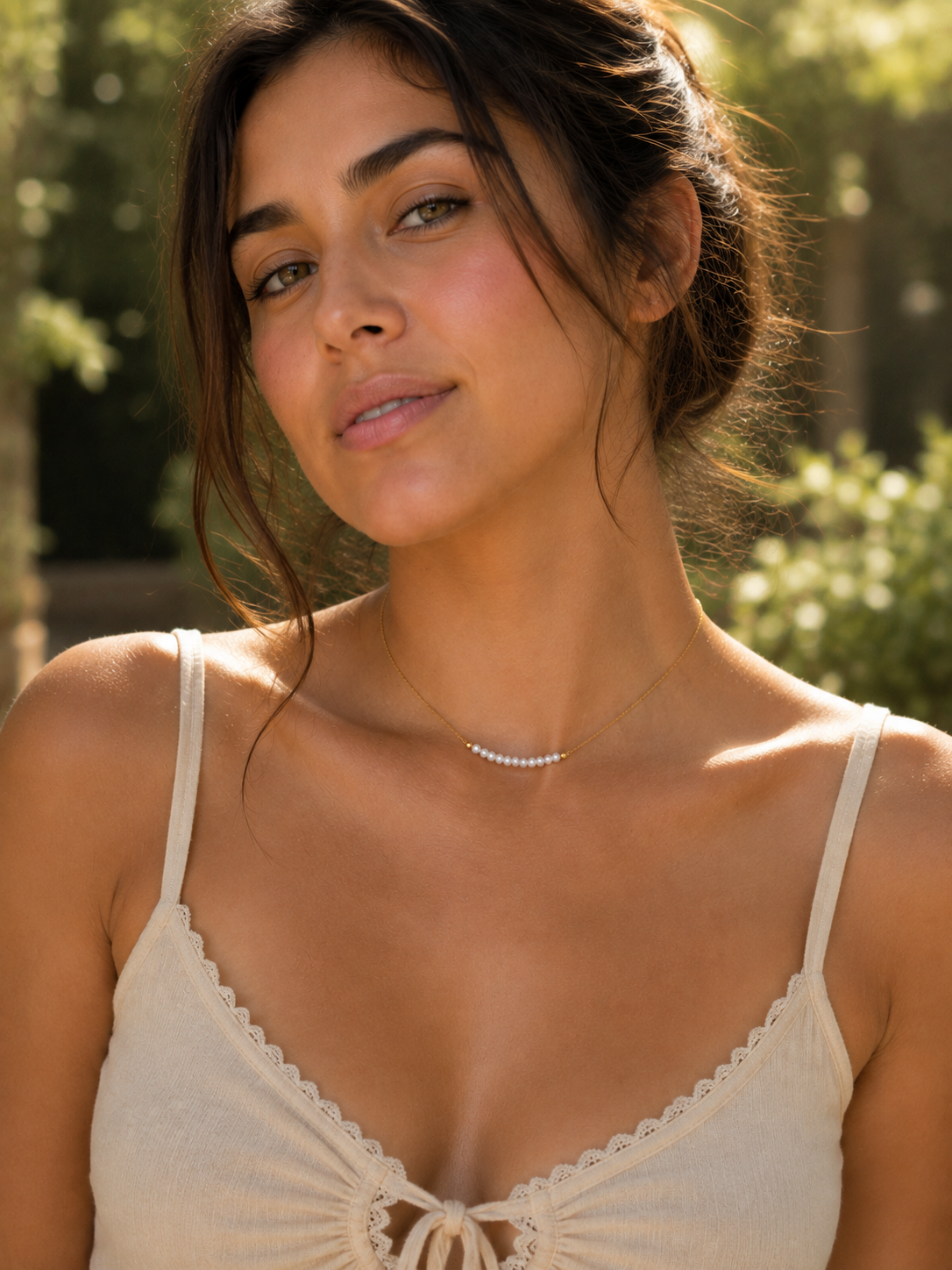 A woman wearing a delicate silver curved necklace with small clear stones, styled with a cream lace-trimmed camisole top, set against a soft-focus outdoor garden background.