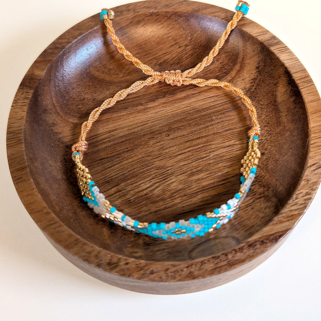 Woven Roots Aqua beaded bracelet with adjustable braided cord, shown in a walnut dish