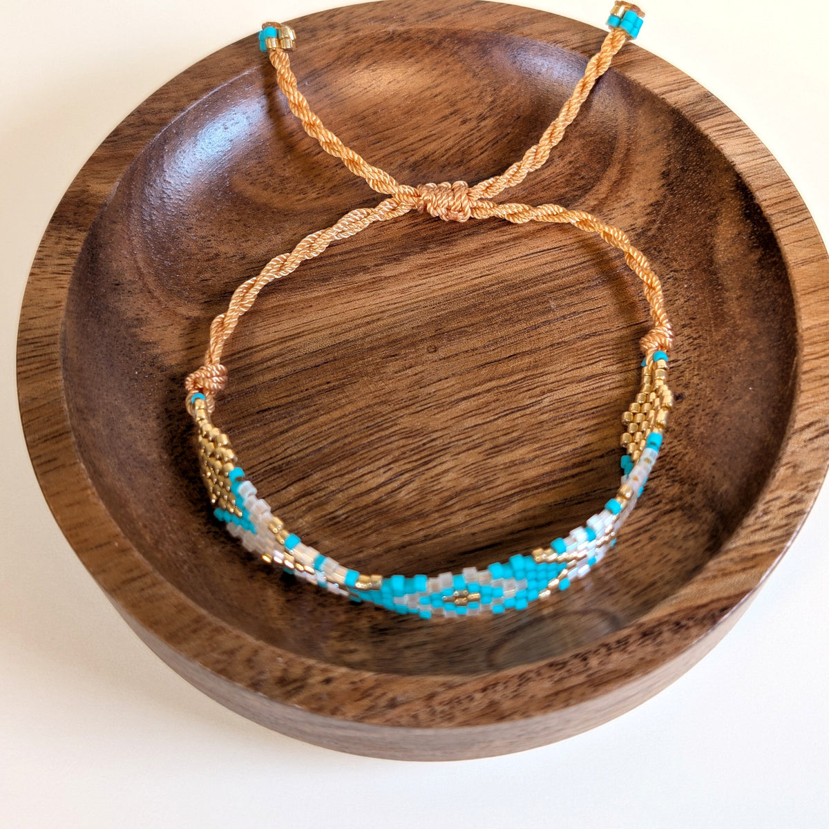 Woven Roots Aqua beaded bracelet with adjustable braided cord, shown in a walnut dish