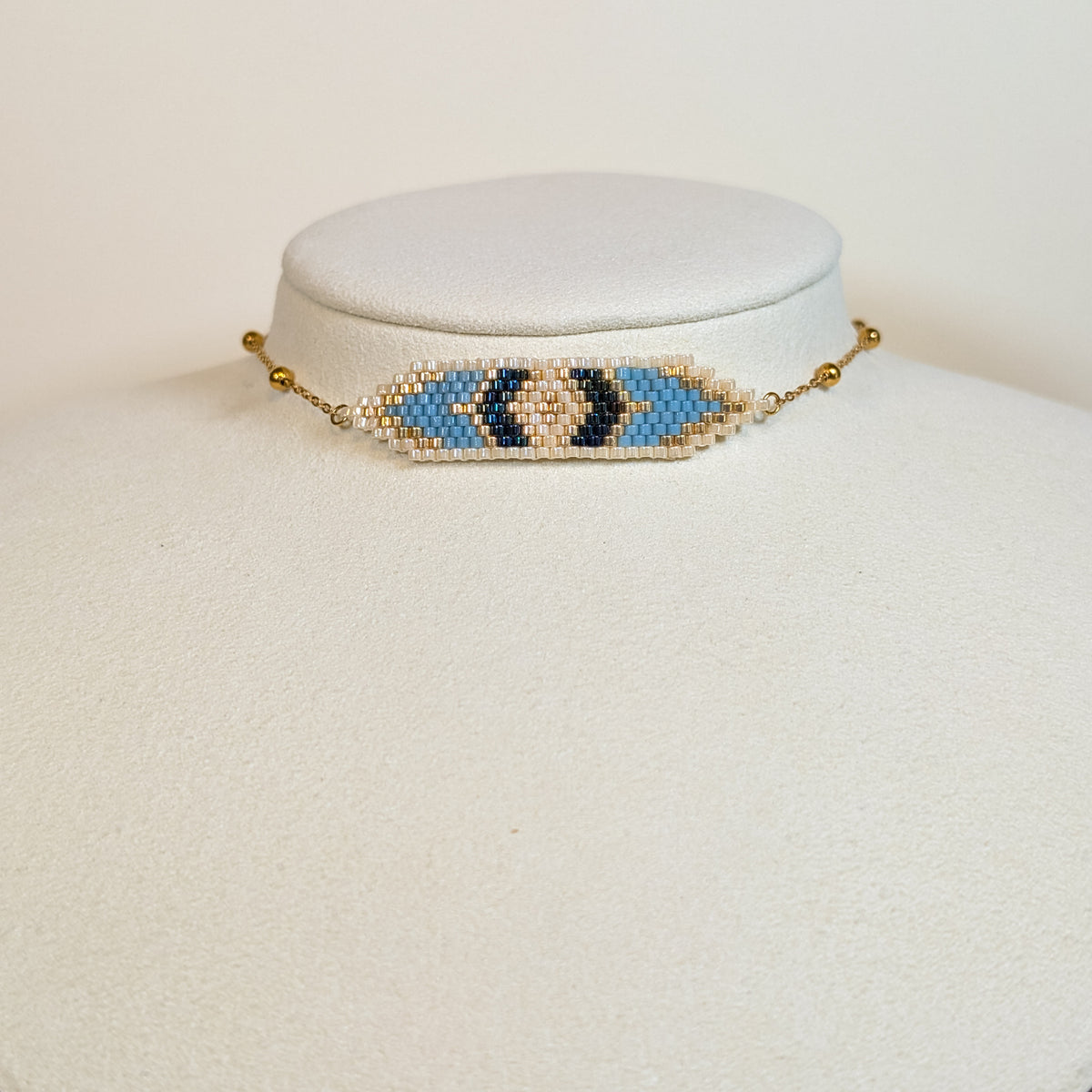 Miyuki beaded choker in pale sky blue and cream with a navy and gold eye motif, on a white display bust