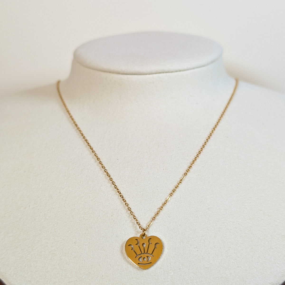 Gold-tone stainless steel necklace with a heart pendant featuring a cut-out eye and crown on a white display bust