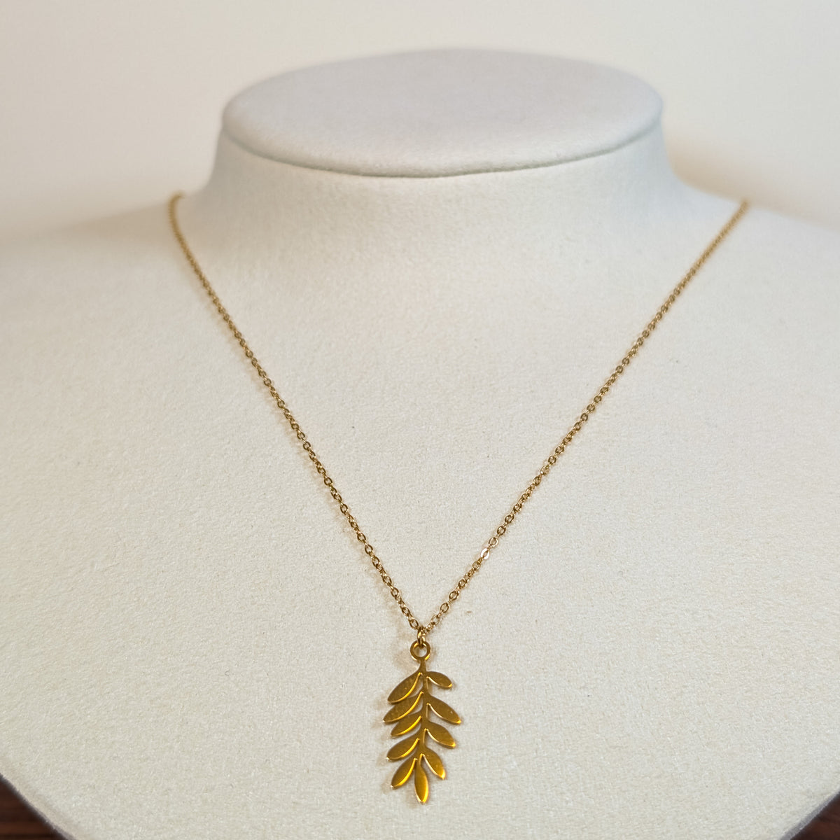 Gold-tone stainless steel necklace with a long fern leaf pendant on a white display bust