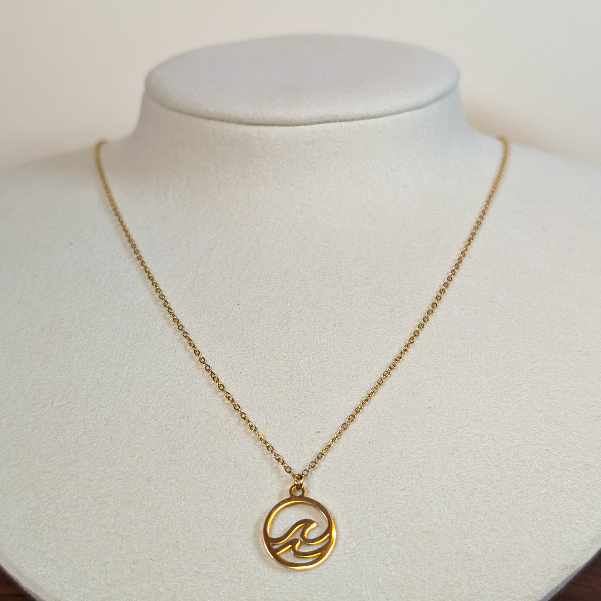 Gold-tone stainless steel necklace with a cut-out wave in a circle pendant on a white display bust